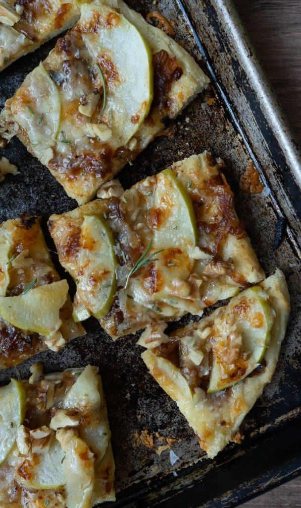 apple and sharp cheddar pizza shows baked pizza with brown bubbly cheese on a sheet pan with an apple and a wood bowl of salt and rosemary sprig in the background