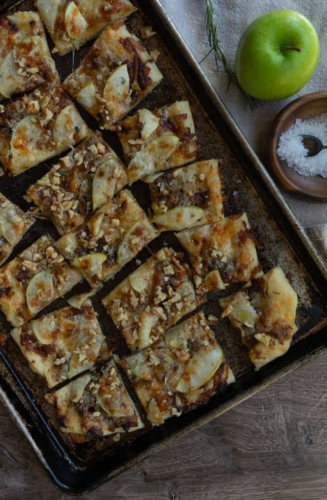 apple and sharp cheddar pizza shows baked pizza with brown bubbly cheese on a sheet pan with an apple and a wood bowl of salt and rosemary sprig in the background