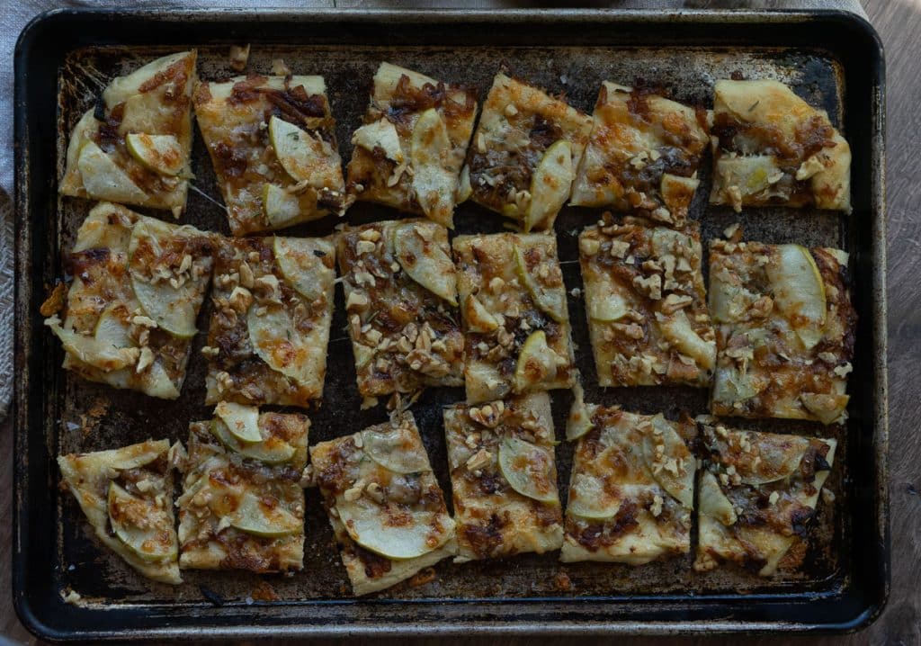apple and sharp cheddar pizza shows baked pizza with brown bubbly cheese on a sheet pan with an apple and a wood bowl of salt and rosemary sprig in the background