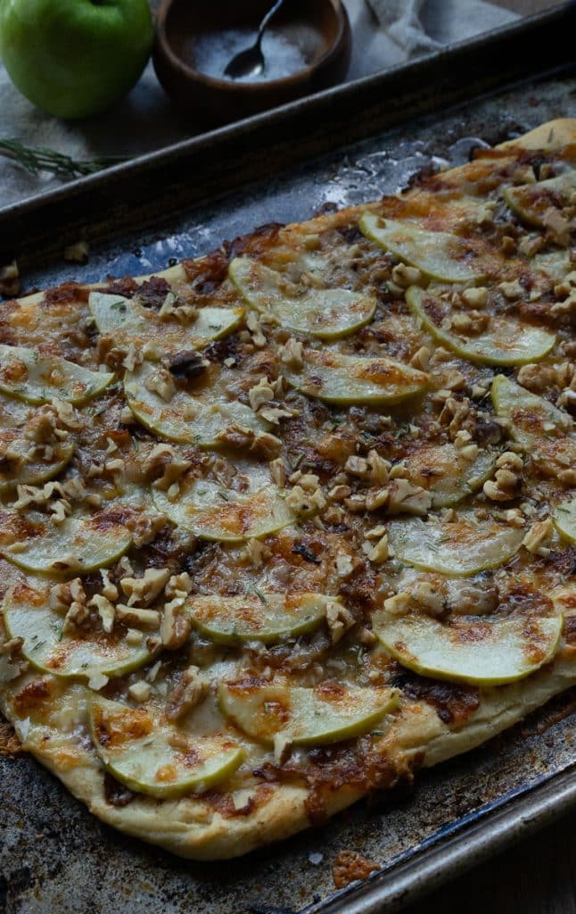 apple and sharp cheddar pizza shows baked pizza with brown bubbly cheese on a sheet pan with an apple and a wood bowl of salt and rosemary sprig in the background