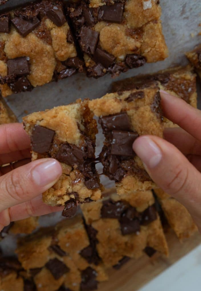 hands pulling apart a salted caramel chocolate chunk cookie bar over the pan of bars