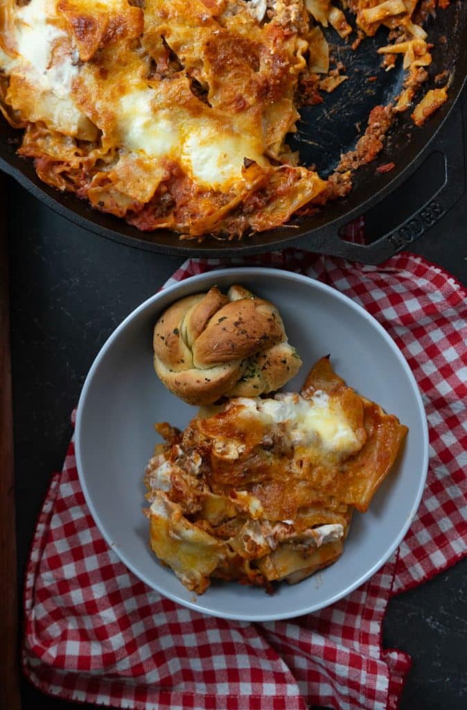 plate of skillet lasagna with a garlic knot on a gray plate with pan of skillet lasagna in the top of the picture