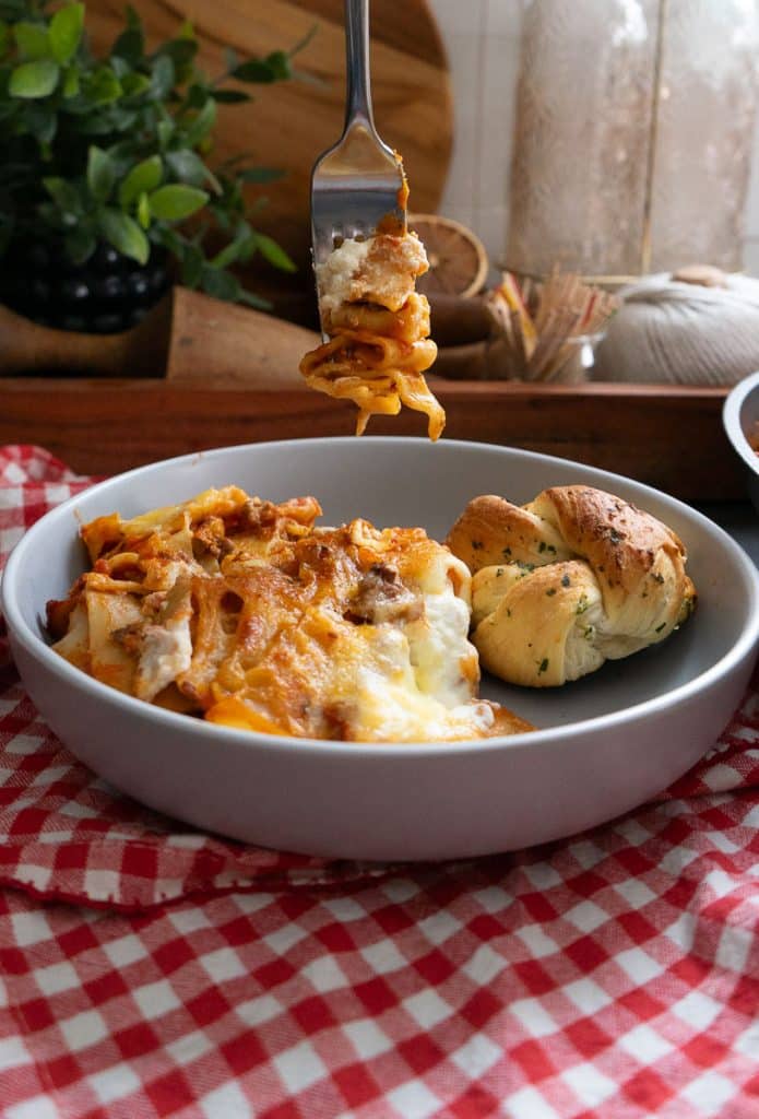fork with lasagna on it hanging over a plate of lasagna on a red and white gingham tablecloth