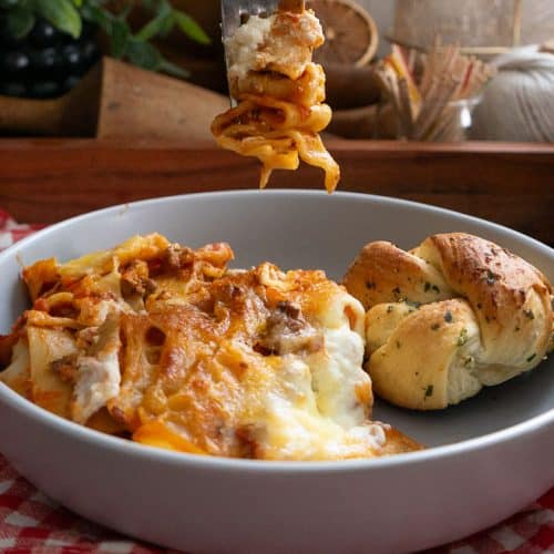 a fork lasagna on it hanging over a plate of skillet lasagna on a red and white gingham tablecloth