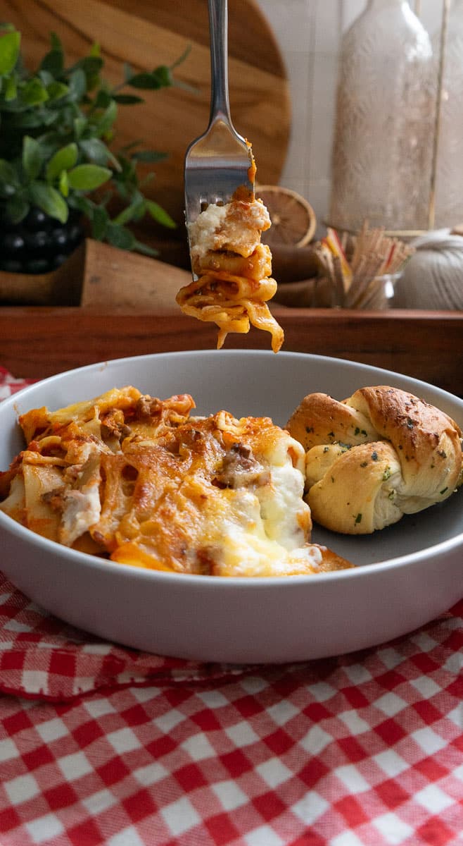 a fork lasagna on it hanging over a plate of skillet lasagna on a red and white gingham tablecloth