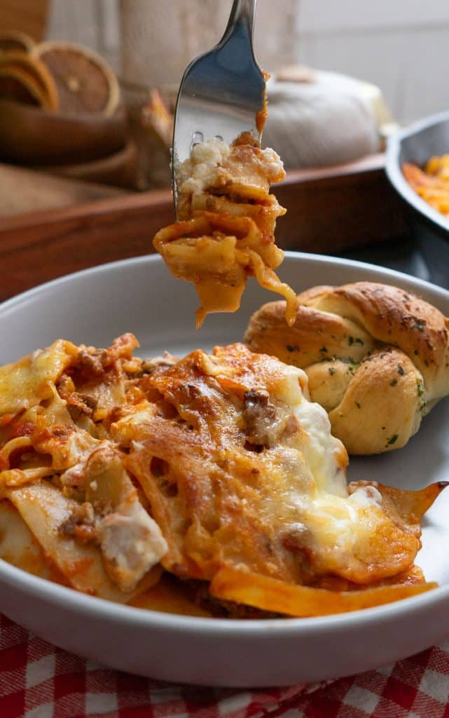 a fork lasagna on it hanging over a plate of skillet lasagna on a red and white gingham tablecloth