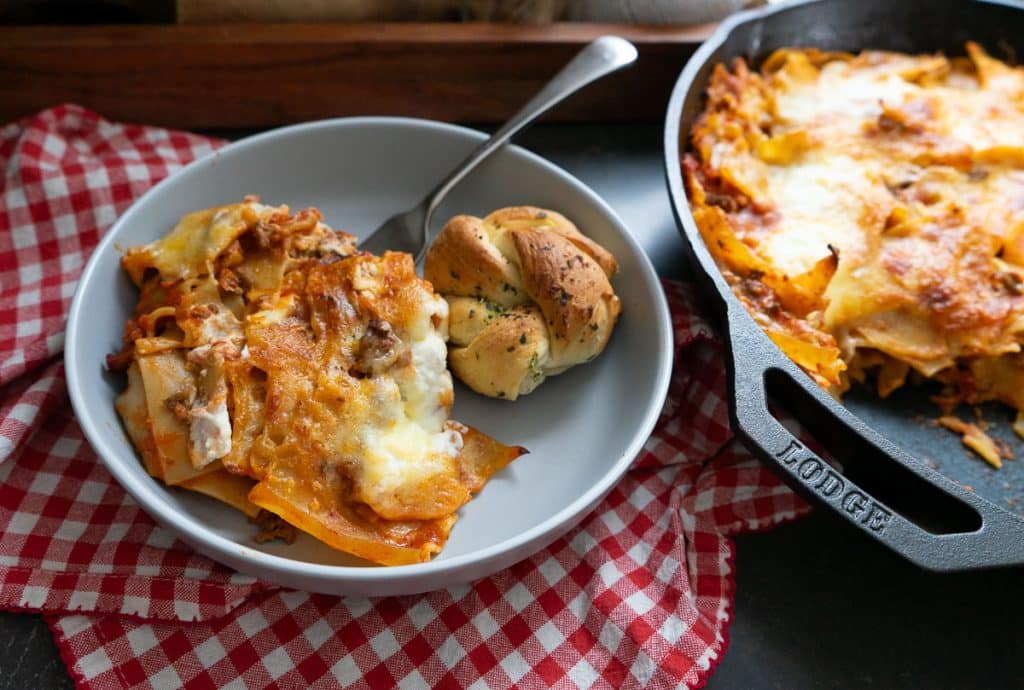 plate of skillet lasagna with a garlic knot on a gray plate sitting next to the pan of skillet lasagna on a red and white gingham tablecloth