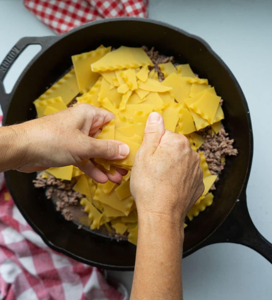hands breaking up lasagna noodles over browned ground beef in a cast iron skillet