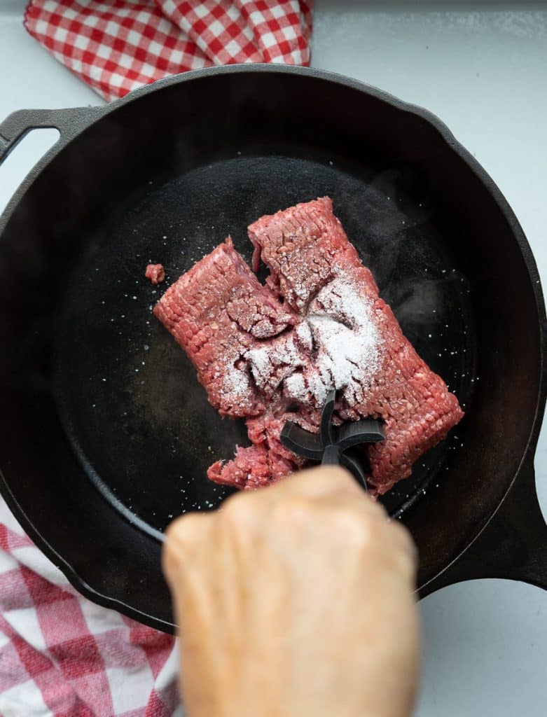 hand breaking up ground beef in a skillet with salt on top of the raw meat in a cast iron skillet