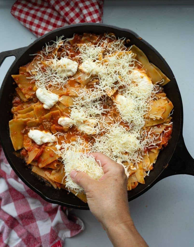 hand sprinkling shredded cheese over the top of the lasagna 