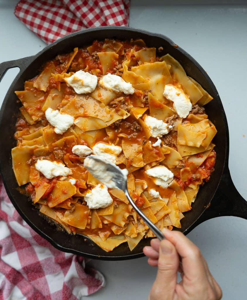 hand with a spoon of ricotta cheese dolloping it over the top of the skillet lasagna