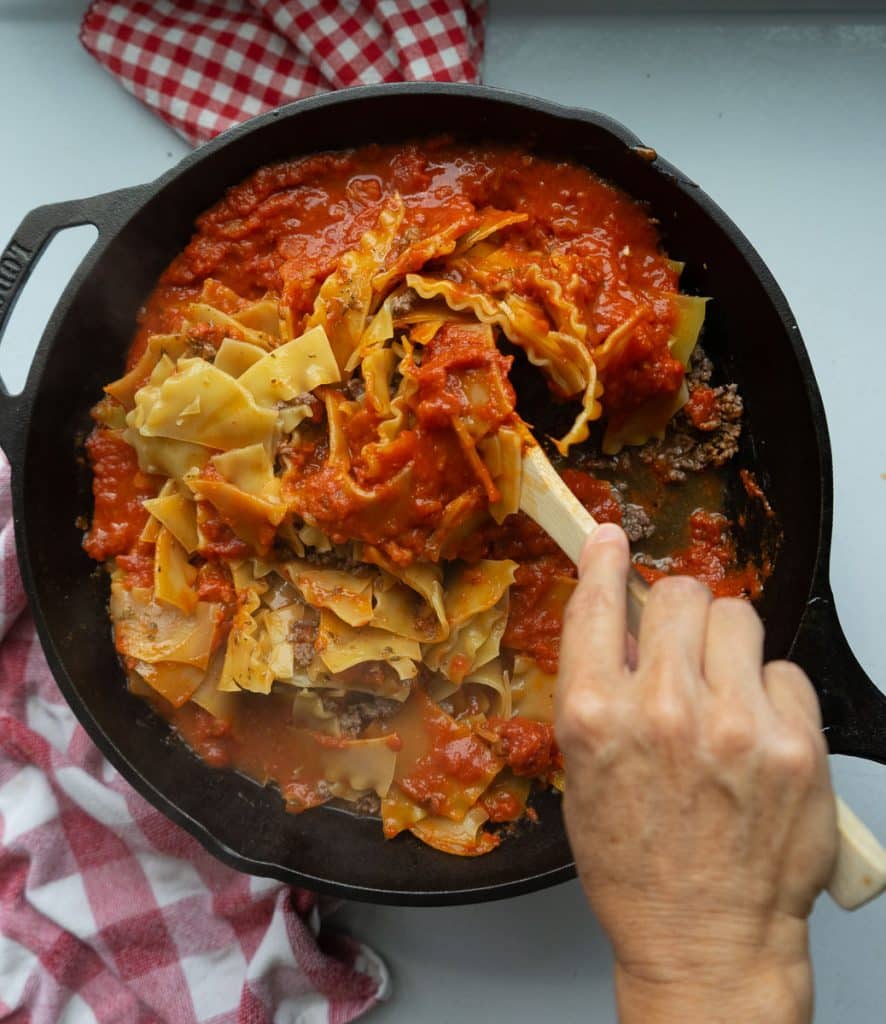 stirring noodles and pasta and meat around with a wooden spatula in a cast iron skillet