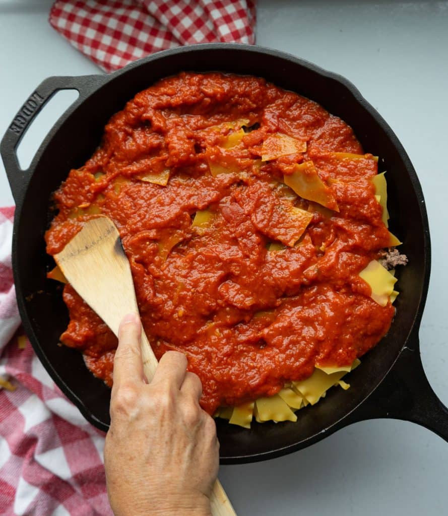 spatula spreading marinara over the noodles in cast iron skillet