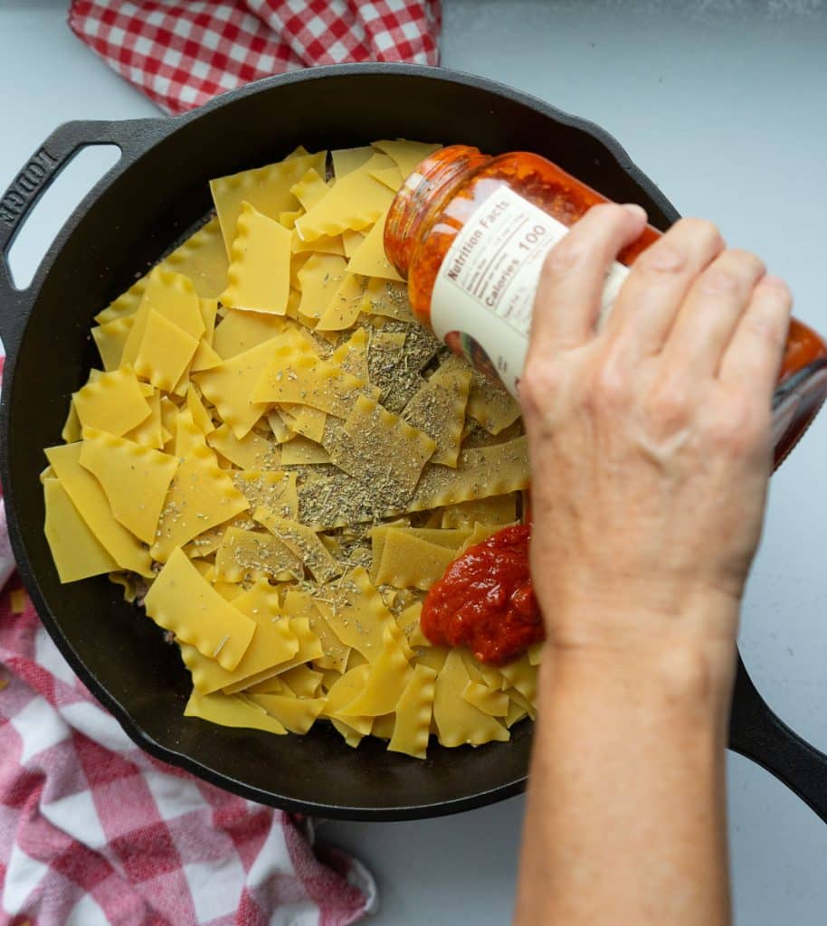 pouring marinara sauce over browned meat and broken noodles in cast iron skillet