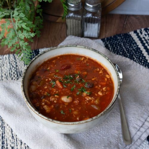 bowl of three bean turkey soup on a tan napkin on a wood table with spoon next to bowl. salt and pepper in back ground with a wood cutting board and a plant