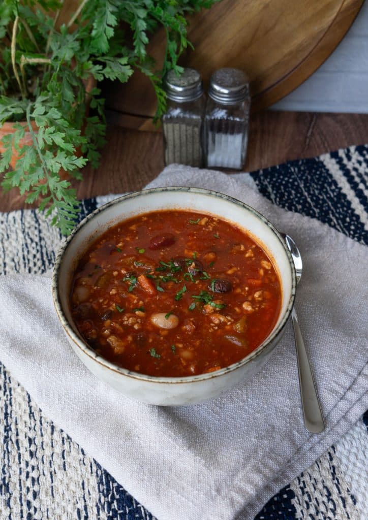 bowl of three bean turkey soup on a tan napkin on a wood table with spoon next to bowl. salt and pepper in back ground with a wood cutting board and a plant