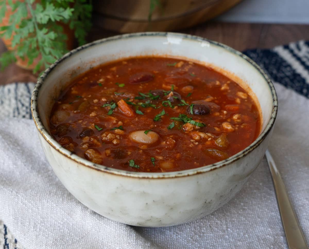 bowl of three bean turkey soup on a tan napkin with a spoon next to the bowl a plant, and a wood cutting board are in the back ground