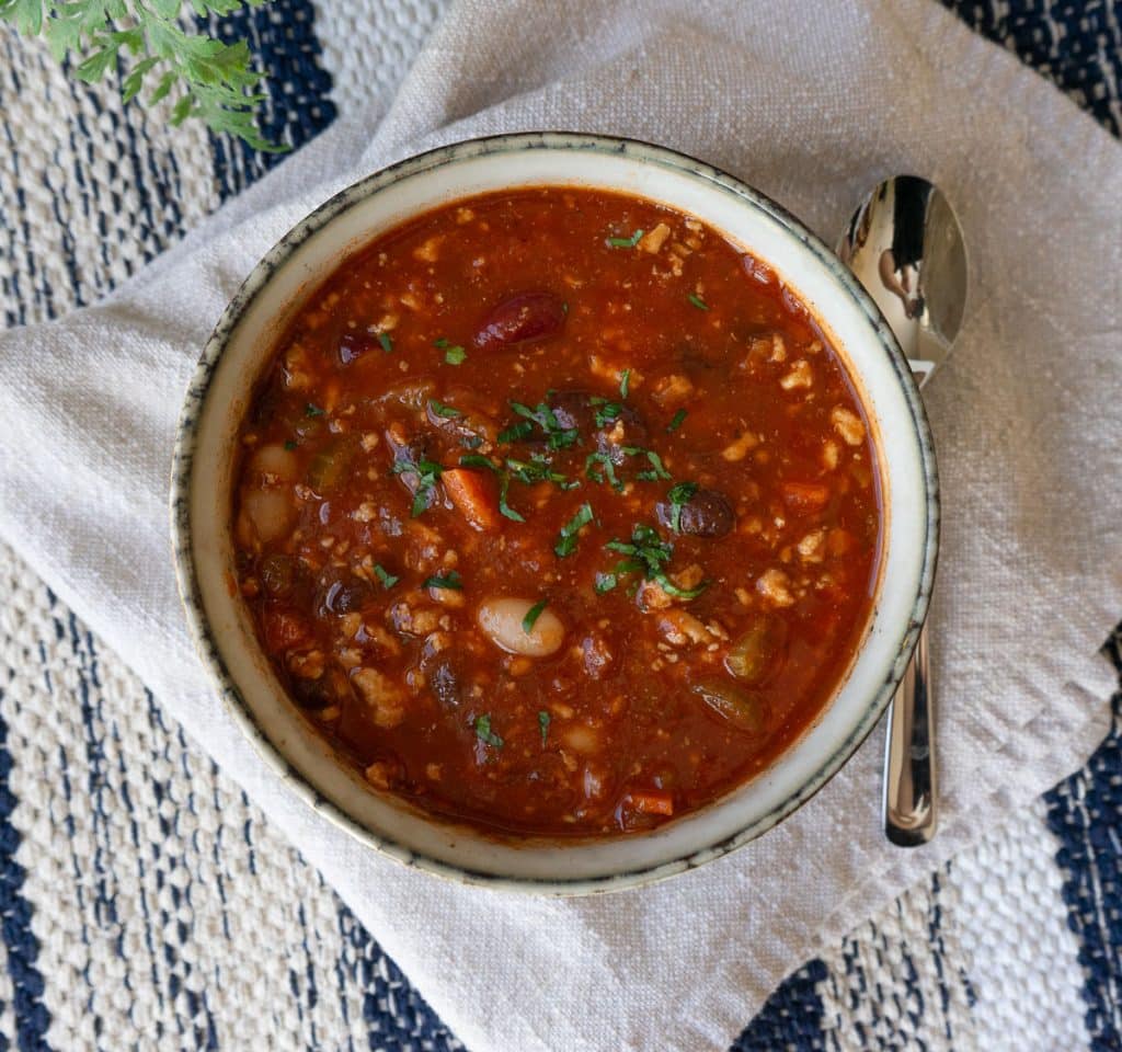 top view of a bowl of three bean turkey soup on a tan napkin with a spoon next to the bowl
