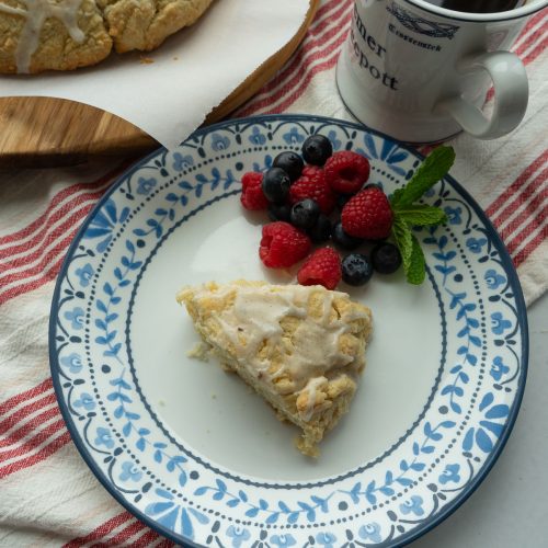 vanilla bean cardamom scone on a plate with berrys on the side, cup of coffee and whole wheel of scones at top of photo