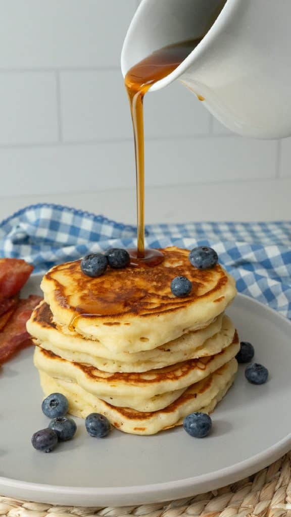 syrup being poured over a stack of pancakaes on a grey plate with blueberries