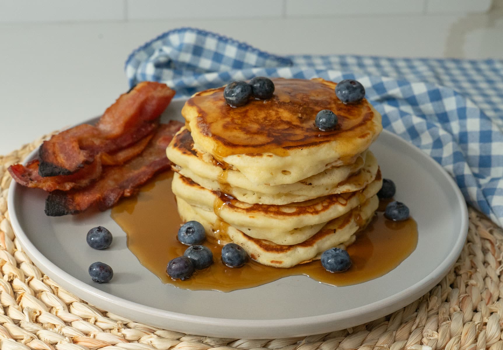Pancakes on a plate covered in maple syrup with blueberries and a side of bacon blue gingham cloth in background