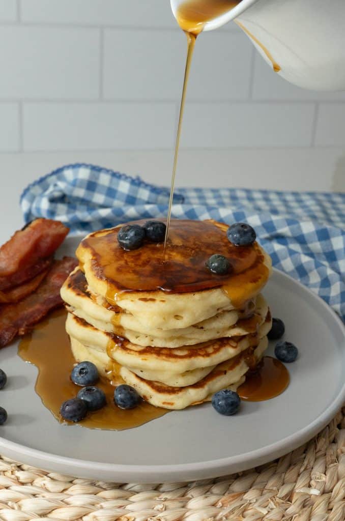 maple syrup being poured over a stack of pancakes with bacon and blueberries on the side