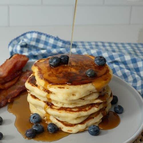 a stack of pancakes getting maple syrup poured over with bacon and blueberries on the side