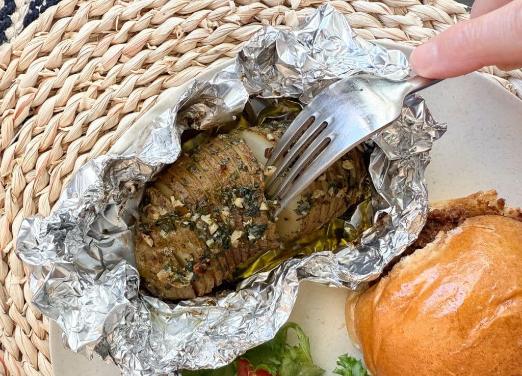 hasselback potato on a plate. the potato is still sitting in the aluminum foil it was baked in, a fork is poking into the potato
