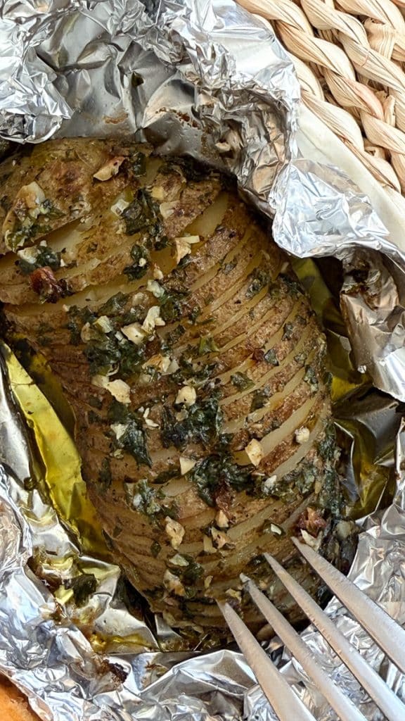 hasselback potato on a plate. the potato is still sitting in the aluminum foil it was baked in, a fork is poking into the potato