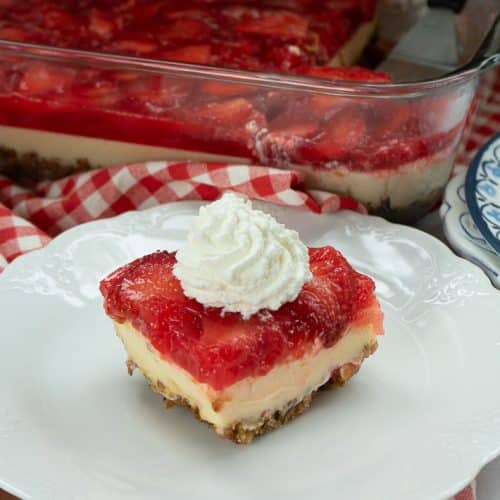 a piece of strawberry cheesecake with pretzel crust on a white plate that is on a red gingham cloth with the whole pan of cheesecake in the back ground with a spatula in the pan