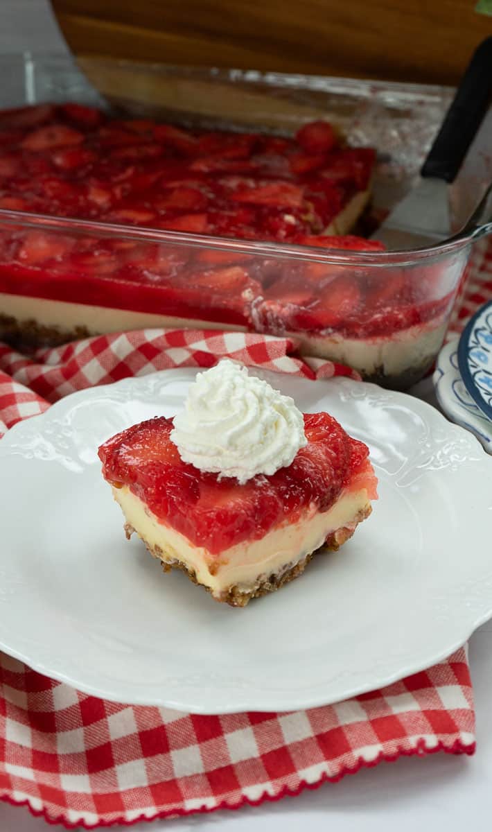 a piece of strawberry cheesecake with pretzel crust on a white plate that is on a red gingham cloth with the whole pan of cheesecake in the back ground with a spatula in the pan