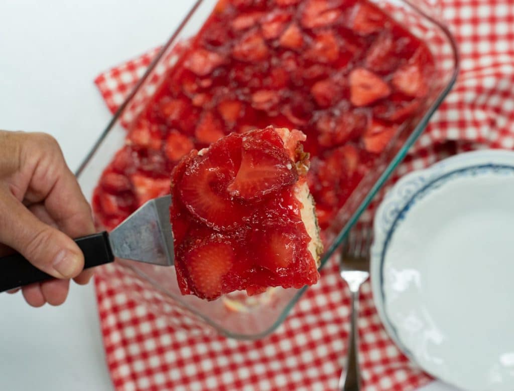 photo of a square of strawberry cheesecake with pretzel crust on a spatula over the dish of cheesecake with plates and a fork on a red gingham cloth