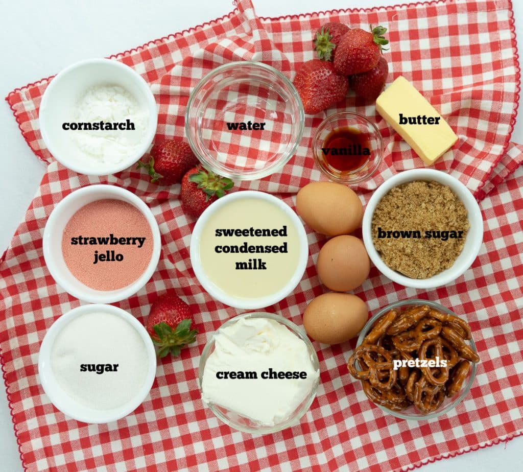 the ingredients for strawberry cheesecake with pretzel crust on a red gingham cloth on a white table