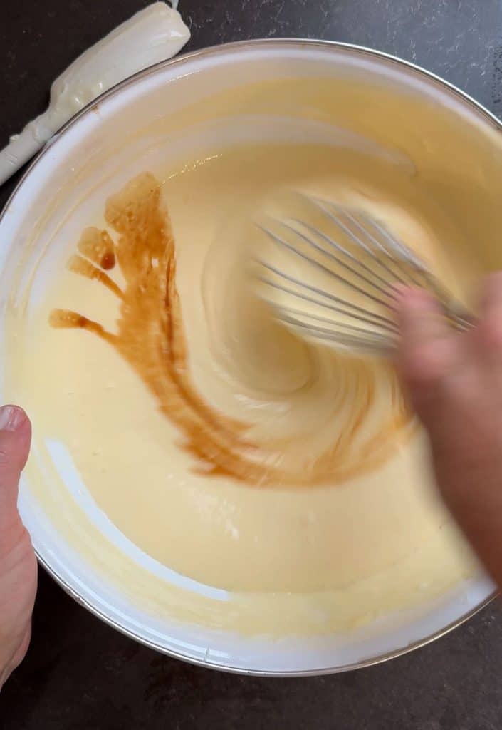 adding vanilla to cheesecake layer in a white bowl on a black counter top silver whisk is stirring