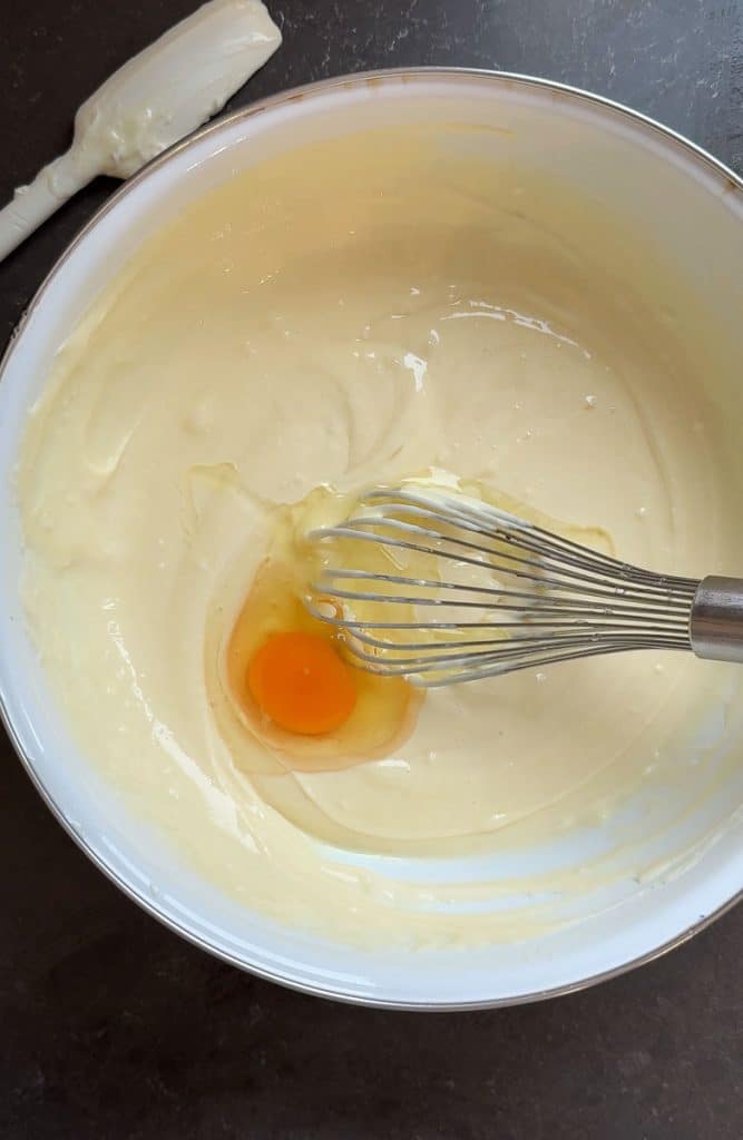 adding eggs to cream cheese cheesecake layer in a white mixing bowl with a silver whisk in the bowl on a black countertop