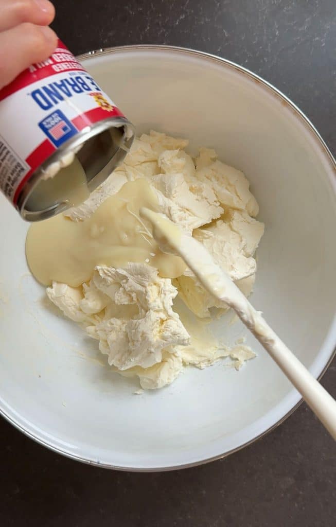 adding sweetened condensed milk to cream cheese in a white bowl on a black countertop