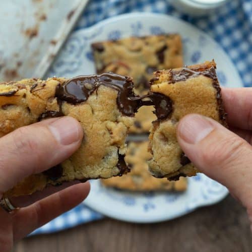 hand pulling apart a cookie bar over plate of caramel pretzel chocolate chip cookie bars on a plate with a glass of white milk next to the whole pan of cut cookie bars
