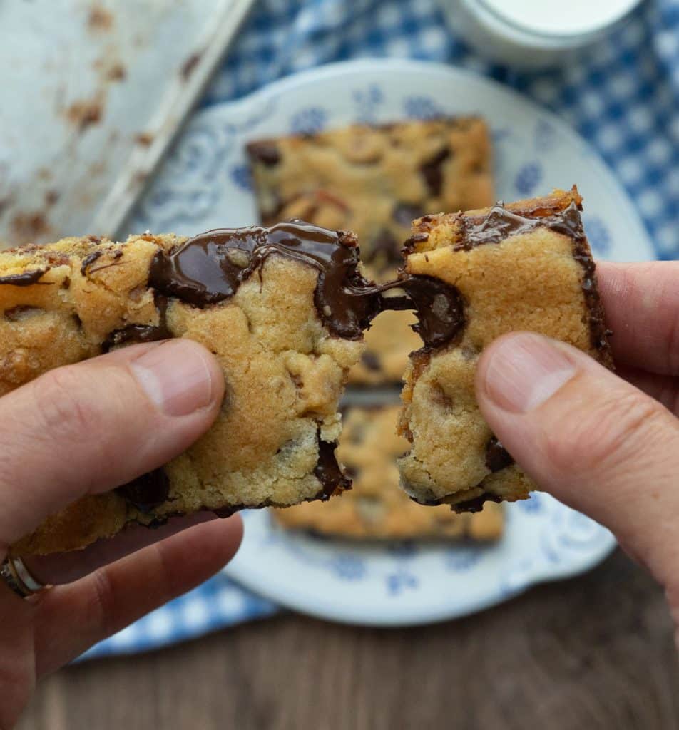 hand pulling apart a cookie bar over plate of caramel pretzel chocolate chip cookie bars on a plate with a glass of white milk next to the whole pan of cut cookie bars