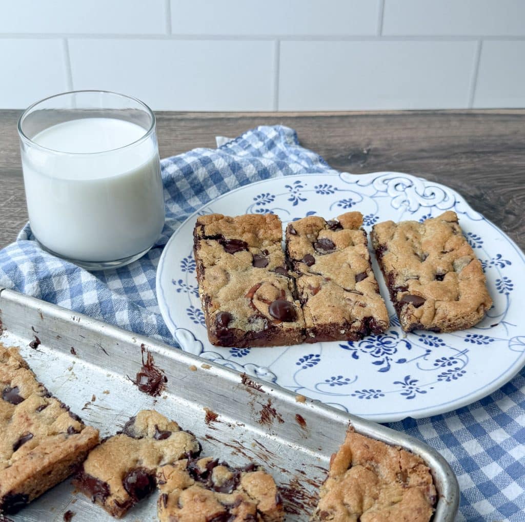 plate of caramel pretzel chocolate chip cookie bars on a plate with a glass of white milk next to the whole pan of cut cookie bars