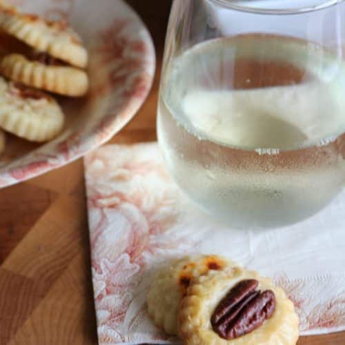 cheese cracker with a pecan on top sitting on a napkin with a glass of white wine in the background and a bowl of cheese crackers are off to the side