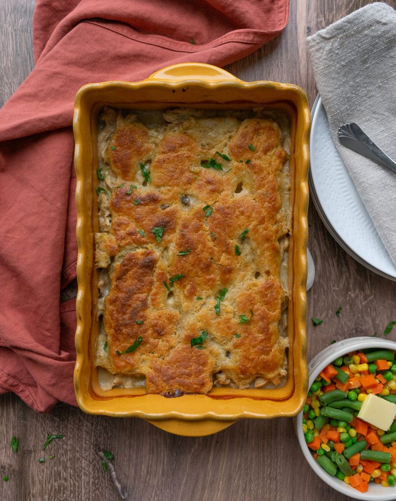 chicken pie baked with a rust colored dish towel off to the left and a bowl of mixed veggies in the lower right hand corner 