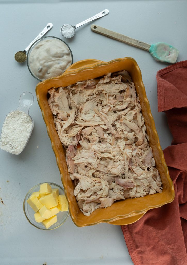 chunks of chicken in casserole dish surrounded by other ingredients in clear dishes a rust colored dish towel is layed to the right of the dish