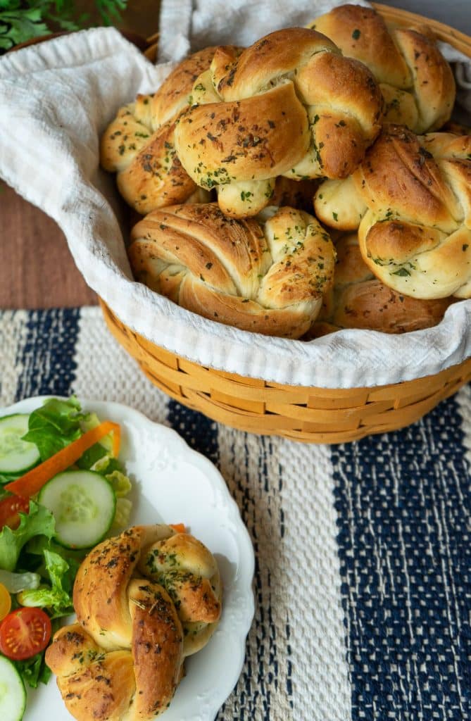 basket of garlic knots next to a plate of salad and a garlic knot on a blue and tan striped placemat