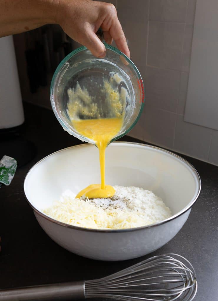 mixing cheese filling together in a white bowl on a black countertop there is a whisk in the foreground and a hand pouring eggs into the bowl