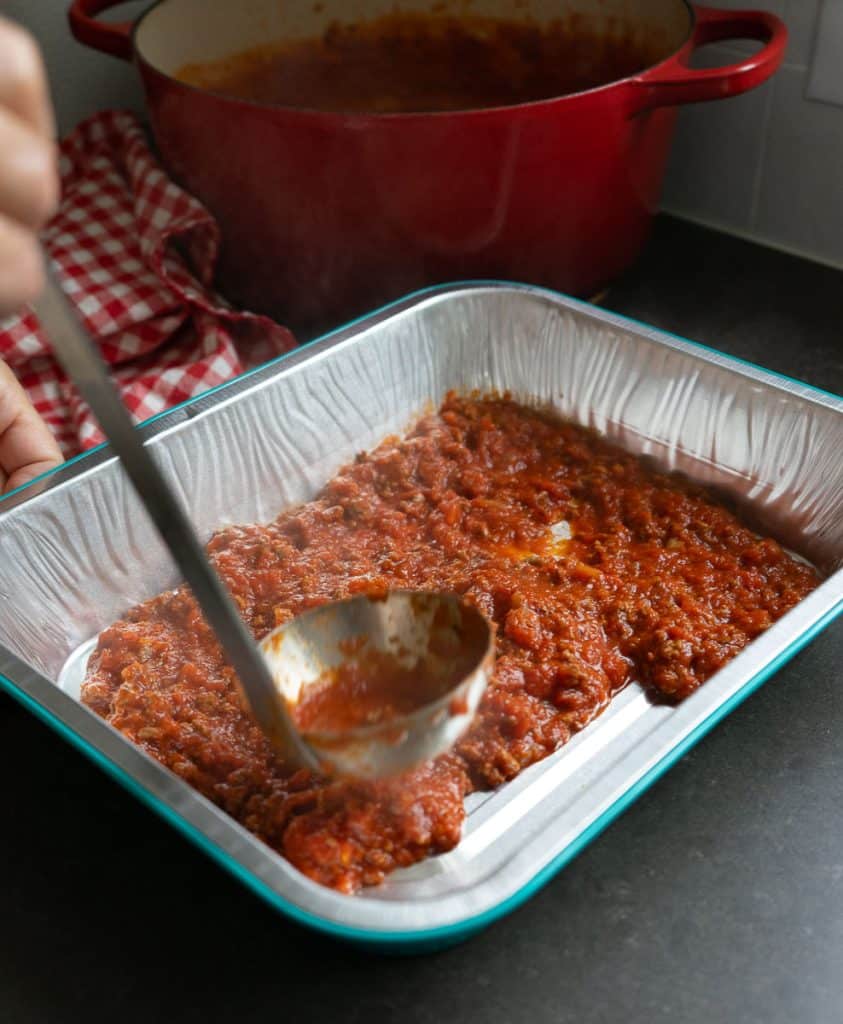 putting meat sauce in the bottom of a 9x13  pan a ladle is spooning sauce into the pan