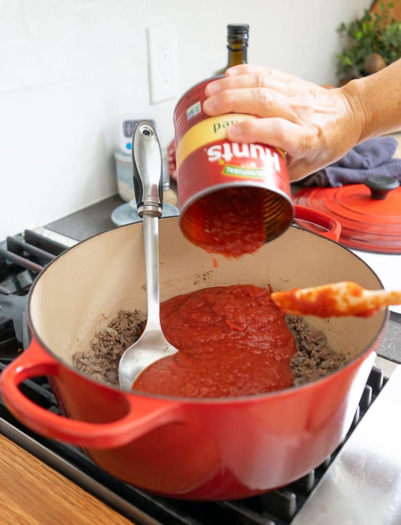 making the sauce in a red pot on the stove top hand is pouring tomatoes into the pot