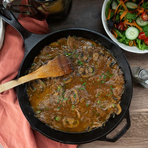 Swiss steak in a cast iron skillet with a wooden spoon on the left side, green salad in a bowl in upper right corner of the photo on a wood counter top