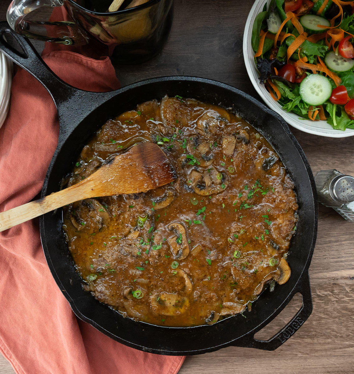 Swiss steak in a cast iron skillet with a wooden spoon on the left side, green salad in a bowl in upper right corner of the photo on a wood counter top