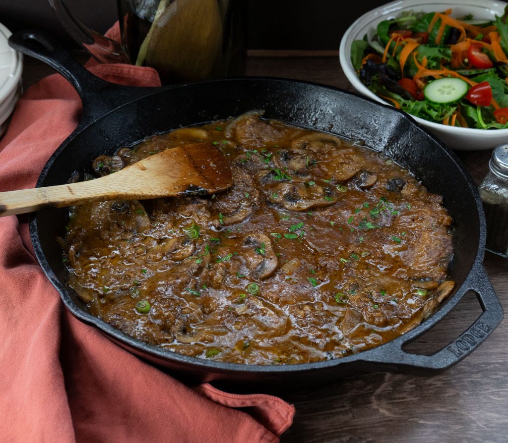 Swiss steak in a cast iron skillet with a wooden spoon on the left side, green salad in a bowl in upper right corner of the photo on a wood counter top