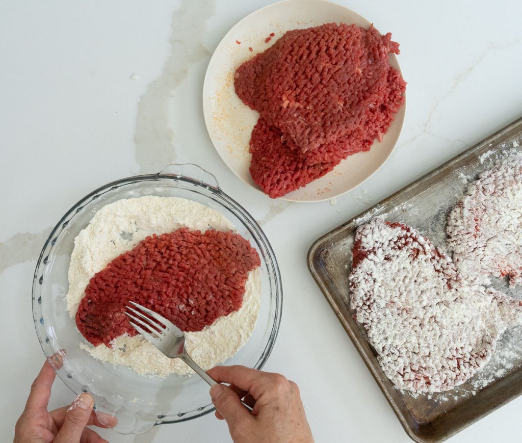 dredging steaks in flour and lawrys season salt mixture. steaks on a plate in upper half of photo and a sheet pan with dredged steaks on it